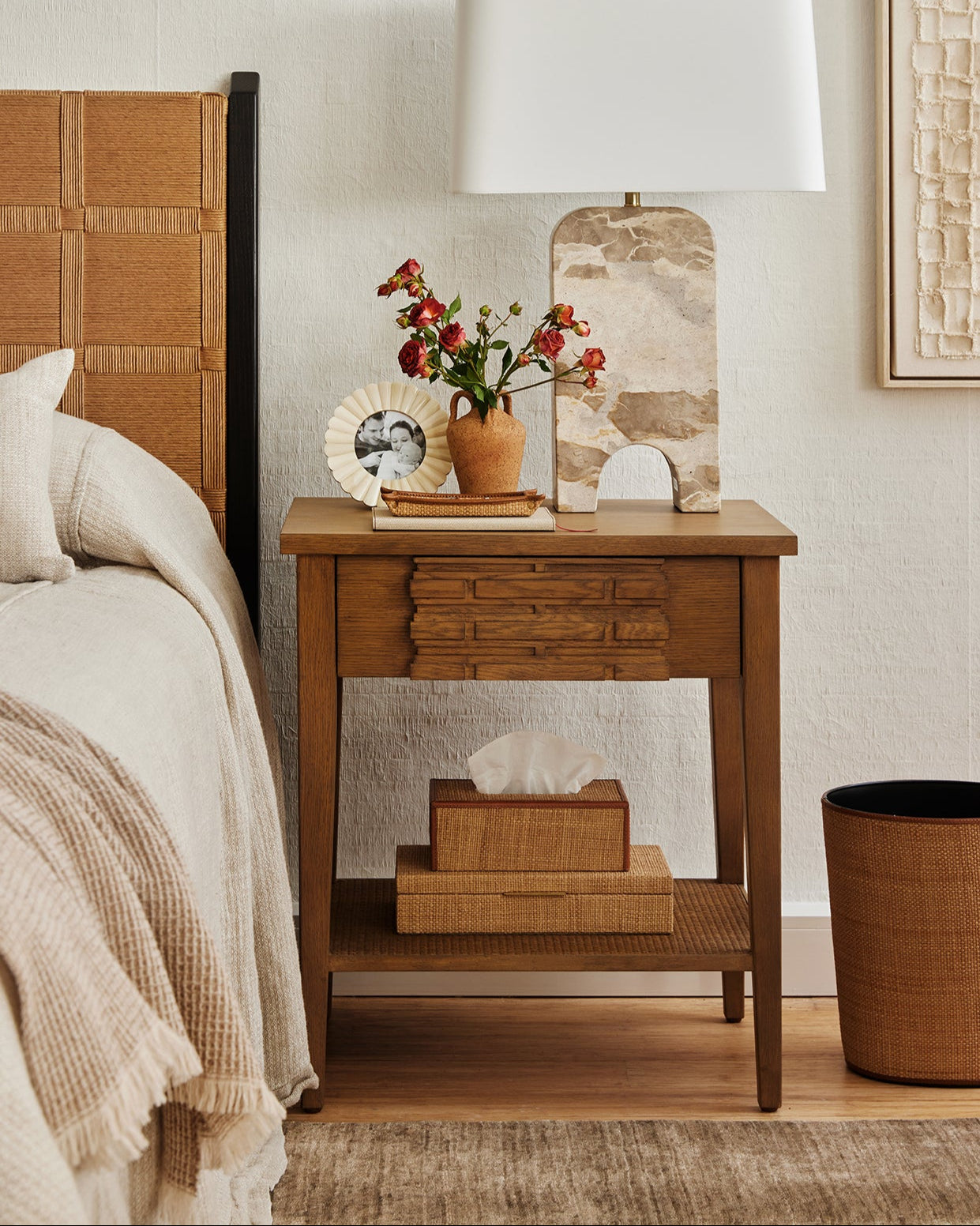 Wooden nightstand with lamp, flowers, and decor items next to a bed.