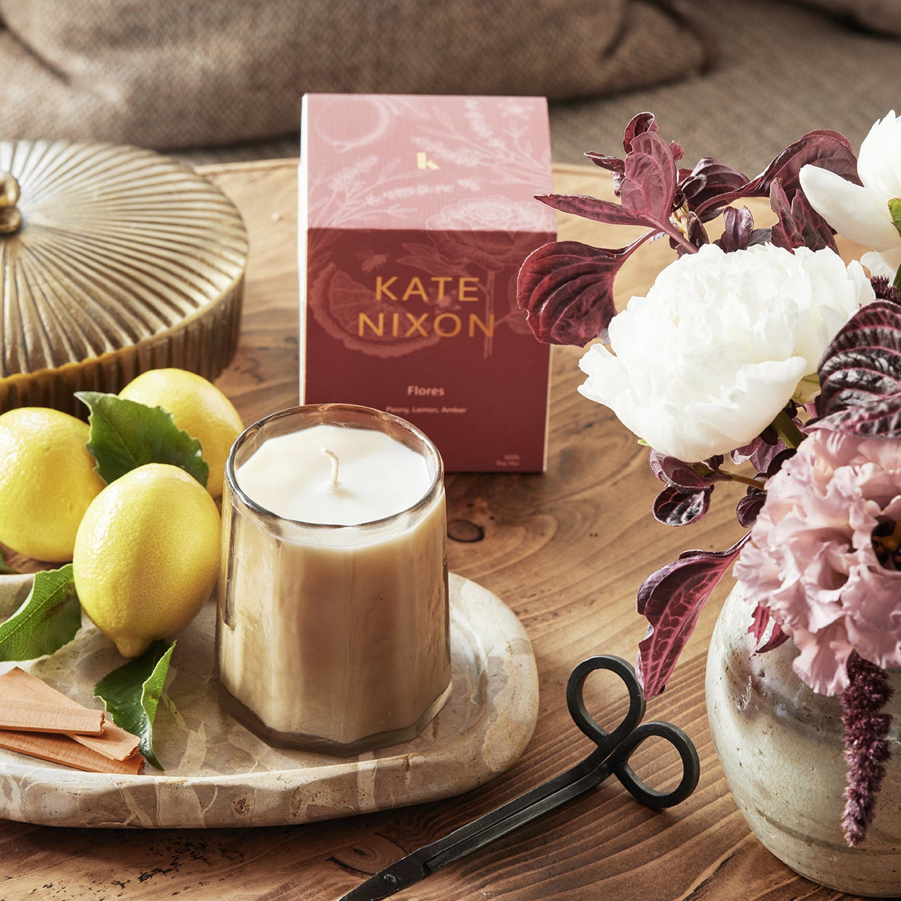 Candle with a decorative box labeled 'Kate Nixon' on a wooden surface with lemons and flowers.