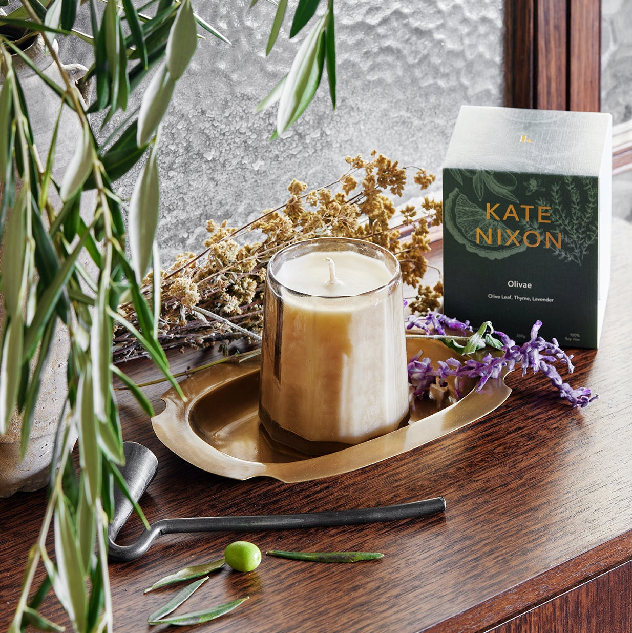 Candle on a gold tray with a box titled 'Kate Nixon' and plants in the background