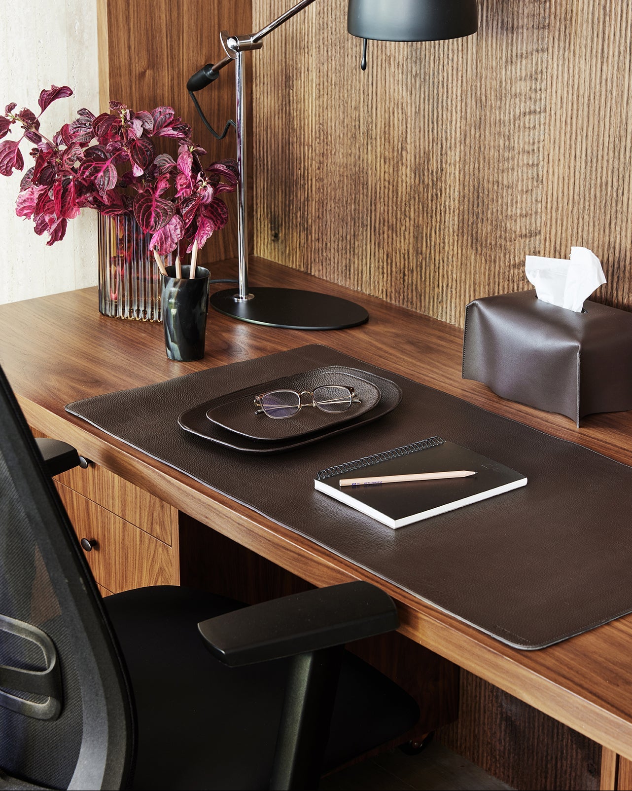 Wooden desk with black lamp, tablet, glasses, and flowers in a room with wooden walls.