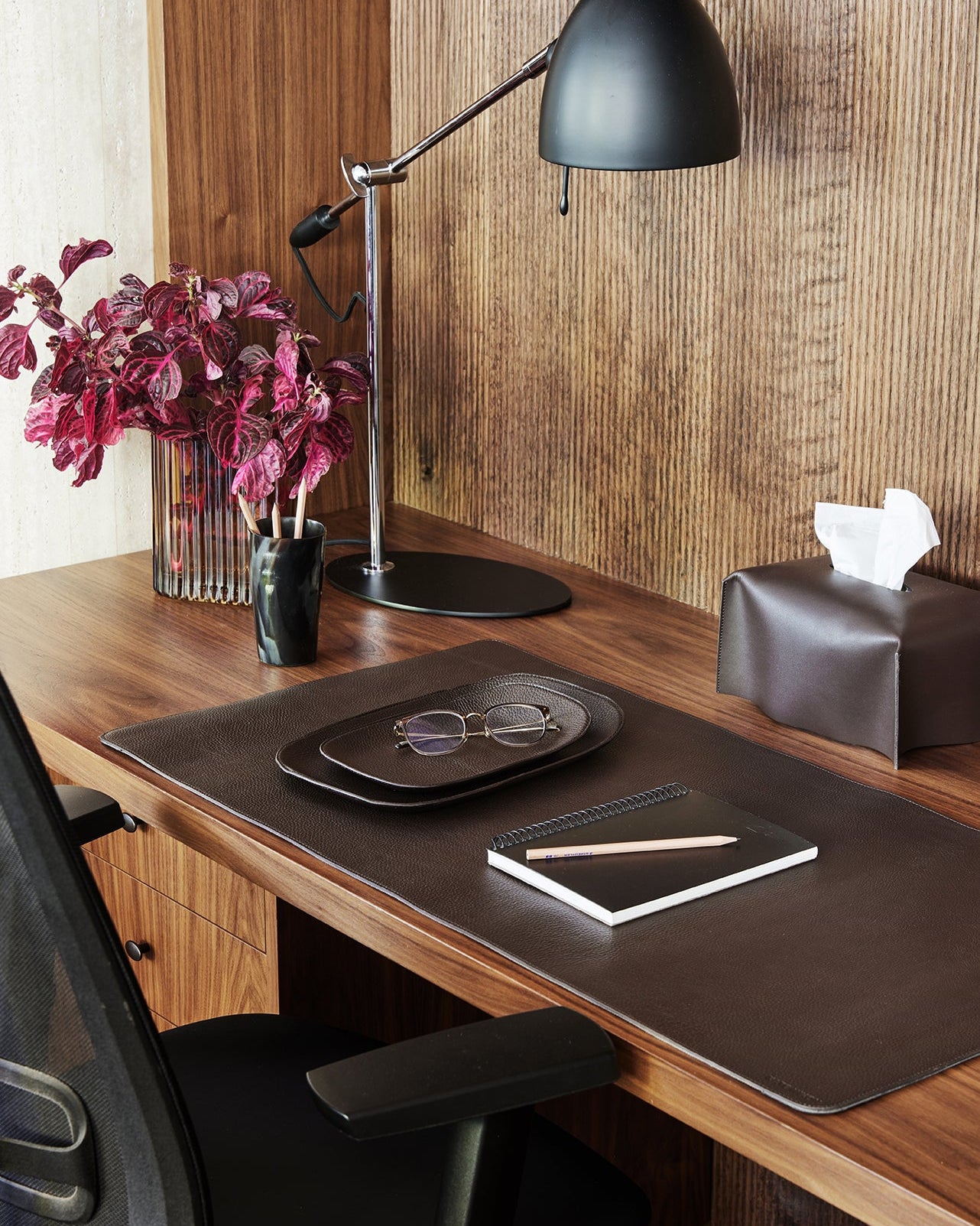 Wooden desk with black lamp, tablet, glasses, and flowers in a room with wooden walls.