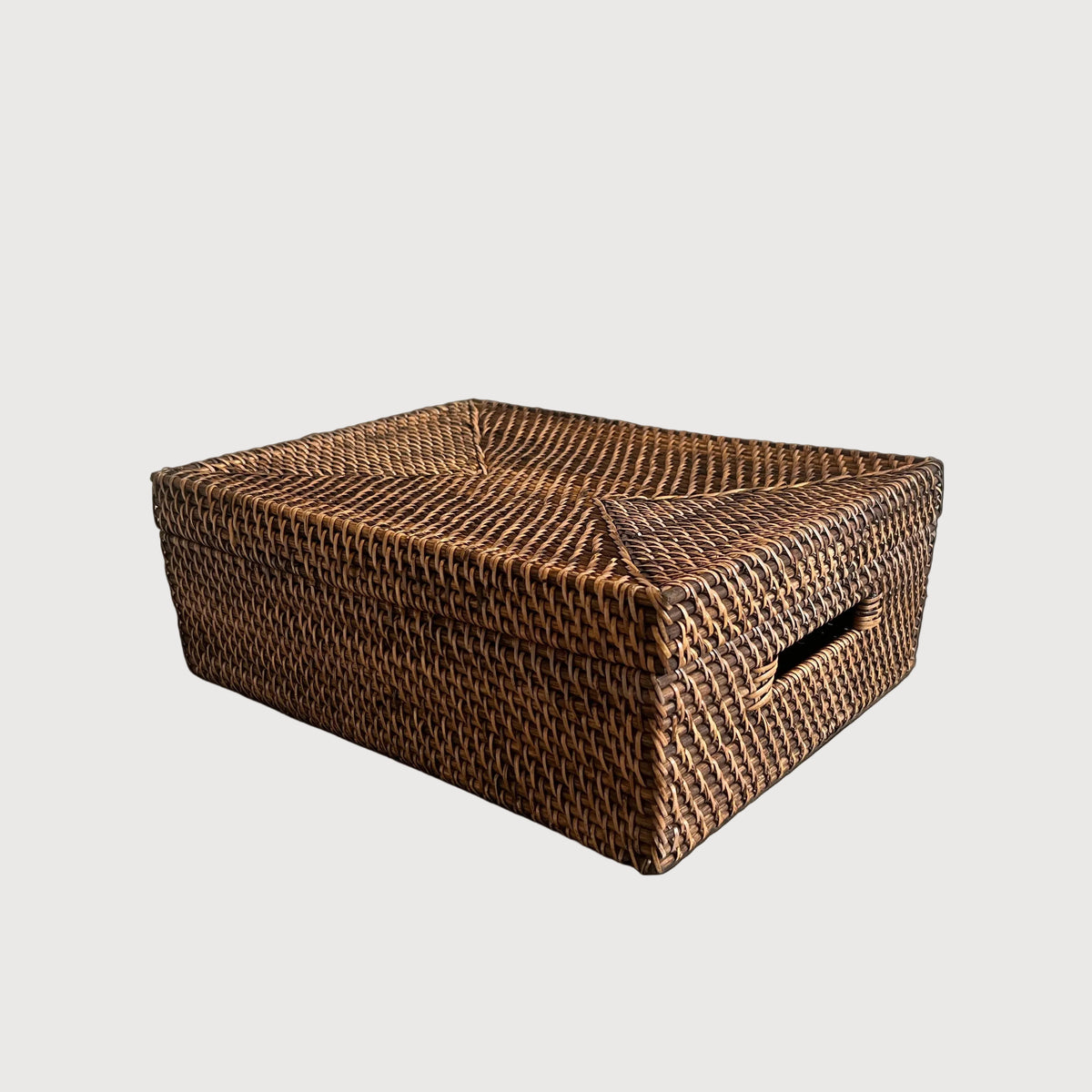 A rectangular brown woven rattan storage box with a lid and a side cutout handle, placed on a stone-tiled floor against a textured gray wall.