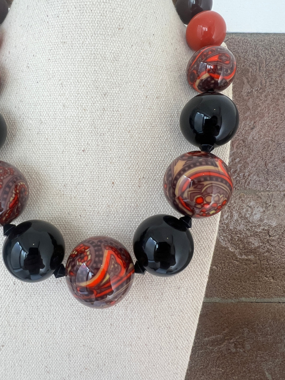 A close-up of a beaded necklace with alternating glossy black round beads and marbled red, orange, and brown patterned beads, displayed on a beige fabric surface.