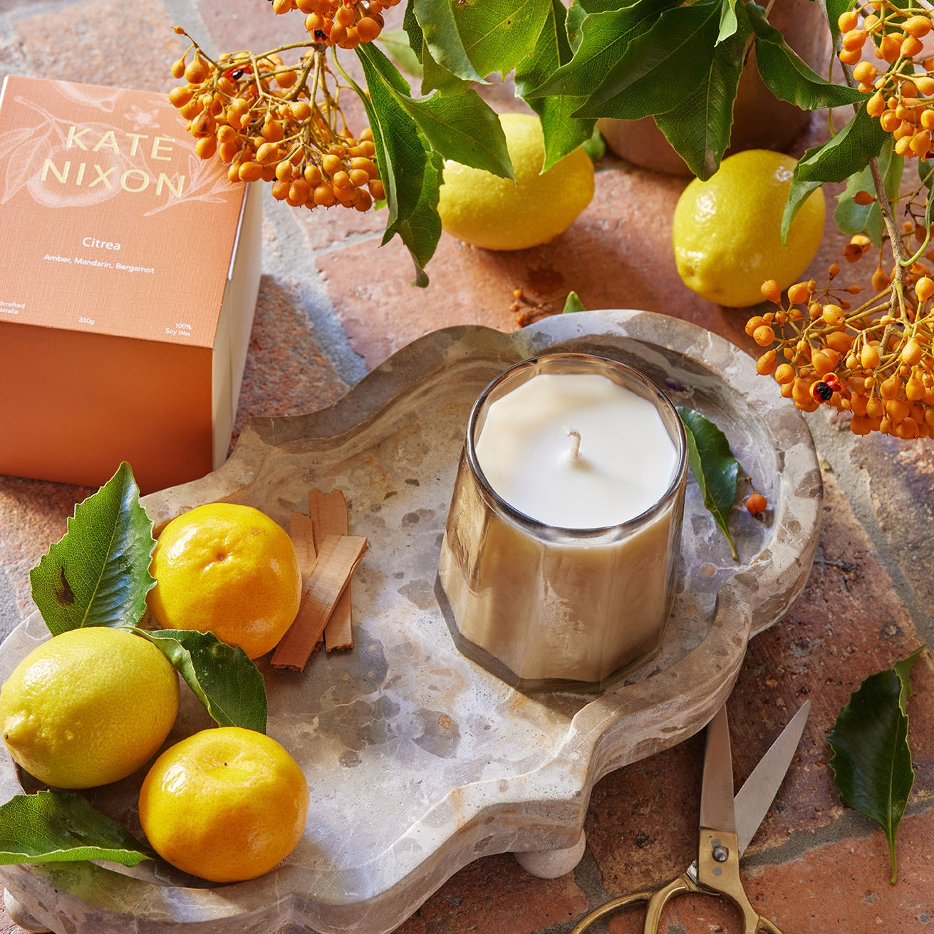 Candle on a decorative tray with lemons, flowers, and scissors on a stone surface