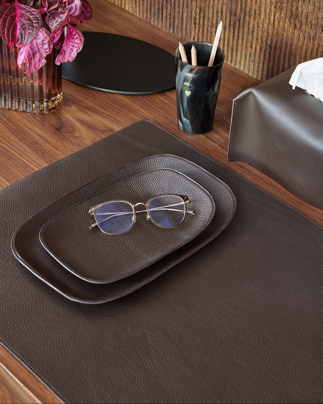 Brown leather tray with sunglasses on a wooden table, next to a vase of pink flowers and a black container.